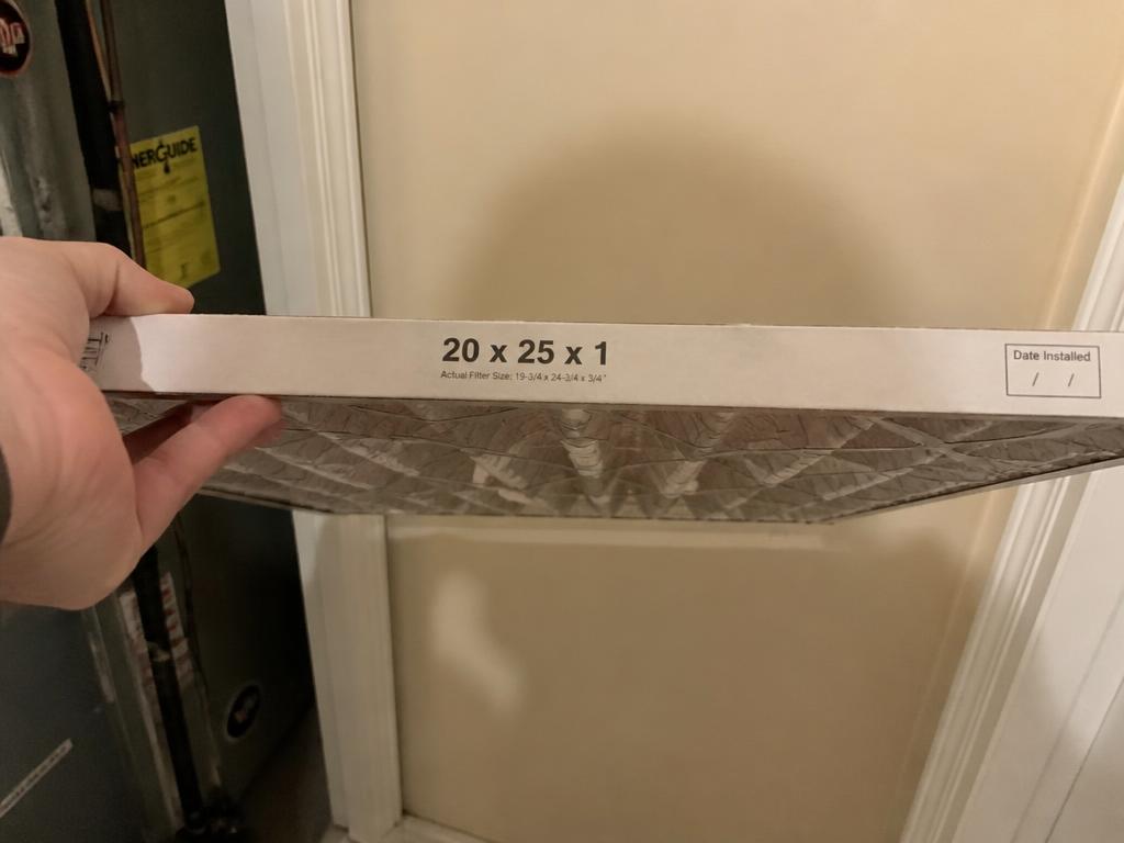A hand holding a household furnace air filter with the printed size visible on the edge